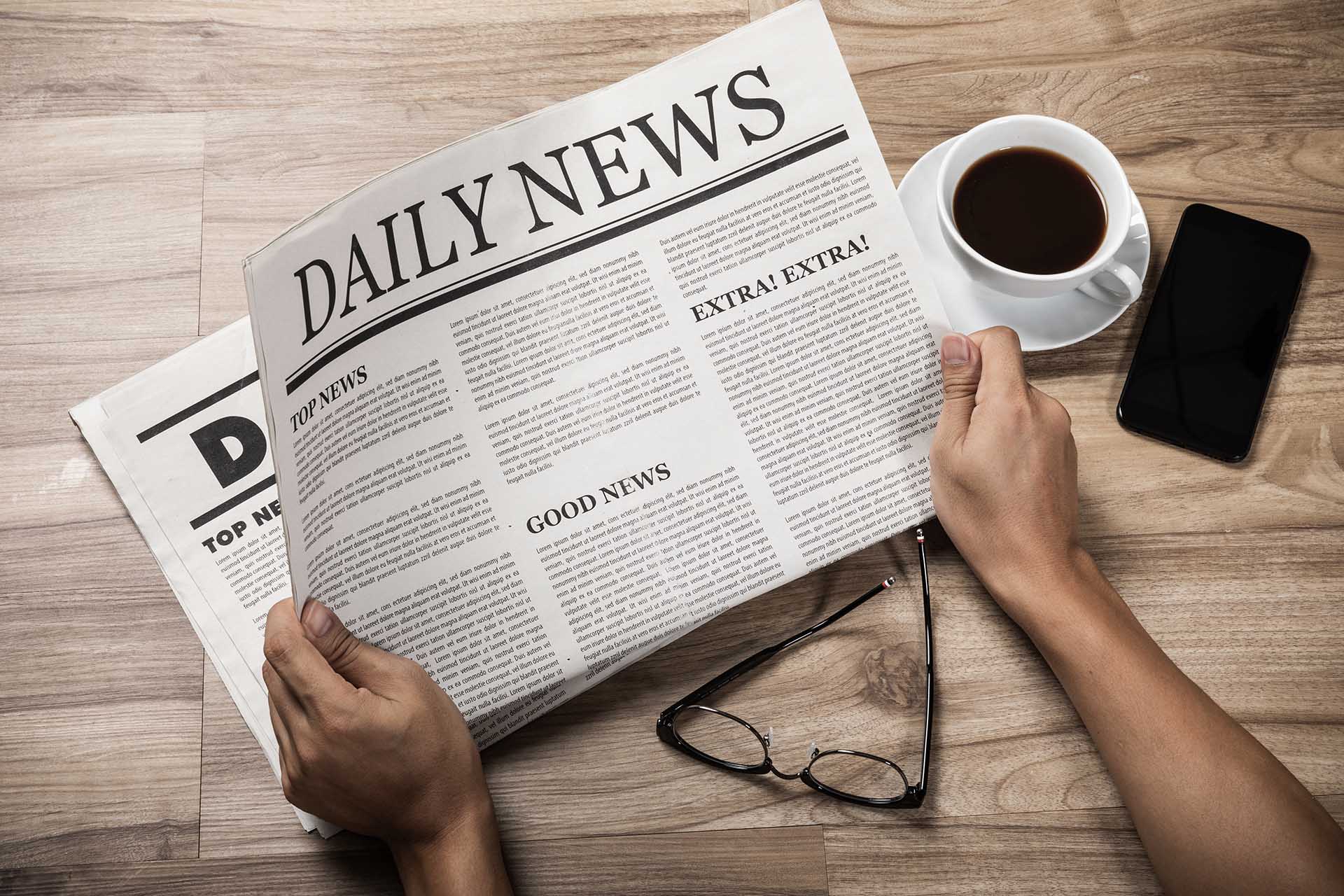 A newspaper showing the daily news page beside a cup of coffee, mobile phone, and eyeglasses.