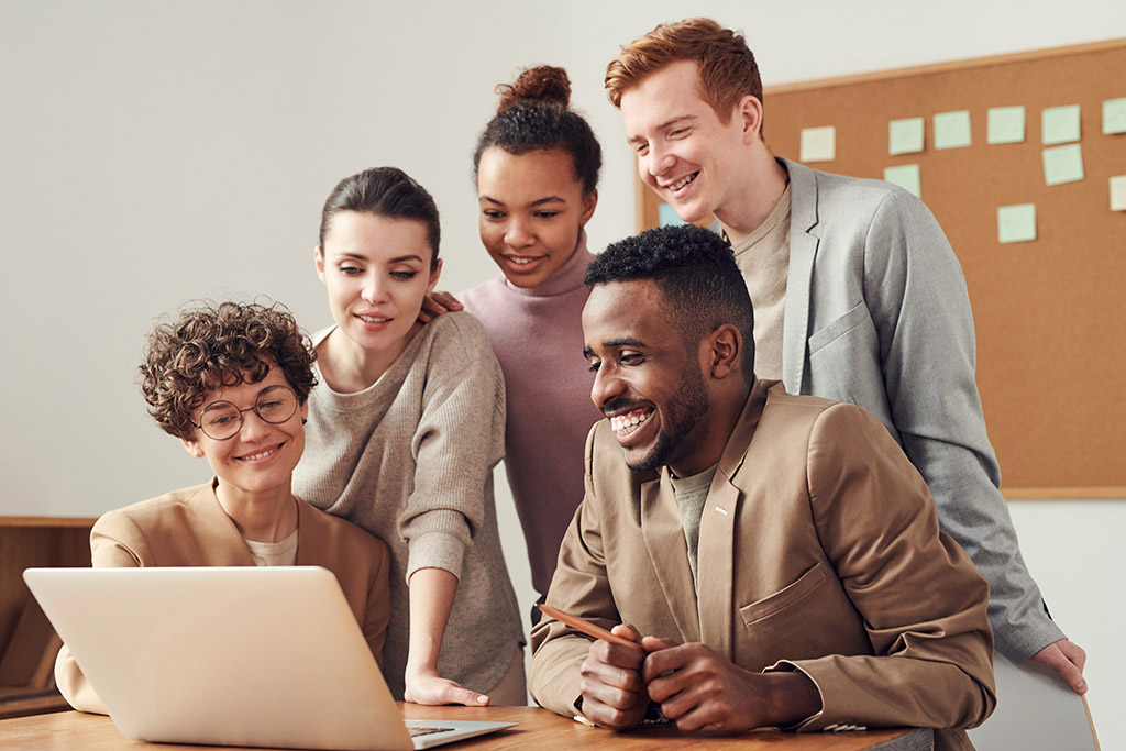 Five smiling people of colors surrounding an open laptop on a table.